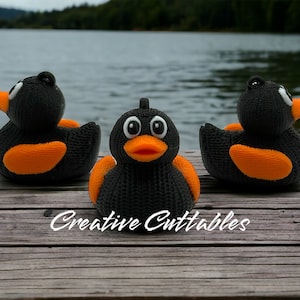 May include: Three black knitted rubber duck toys with orange beaks and feet. The ducks are sitting on a wooden surface. The ducks are facing different directions. The text "Creative Cuttables" is visible in the bottom center of the image.