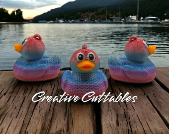 May include: Three rubber duck toys with a rainbow ombre color scheme. The ducks are sitting on a wooden dock with a lake in the background. The text "Creative Cuttables" is visible on the dock.