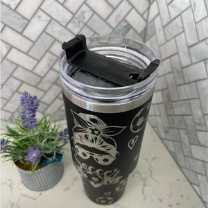May include: Black stainless steel tumbler with a black and white design of a woman wearing a bandana and sunglasses with the words "Soccer Mom" and soccer balls.