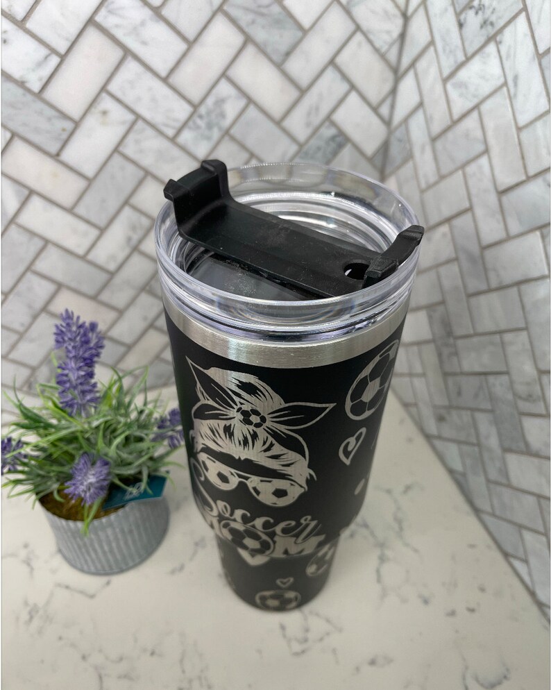 May include: Black stainless steel tumbler with a black and white design of a woman wearing a bandana and sunglasses with the words "Soccer Mom" and soccer balls.