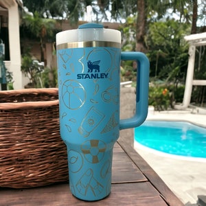 May include: A blue stainless steel travel mug with a gold Stanley logo and a gold design of beach-themed items. The mug has a lid with a straw.