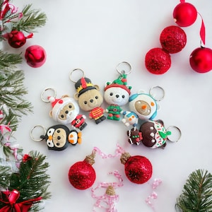 May include: A set of seven Christmas-themed keychains featuring various characters, including a penguin, a snowman, a gingerbread man, and a bear. The keychains are arranged in a circle on a white background with red and white Christmas ornaments and ribbon.