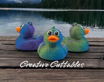 May include: Three rubber duck toys with a textured, knitted-like surface. The ducks are blue, green, and purple. The ducks are sitting on a wooden surface. The text "Creative Cuttables" is visible in the bottom center of the image.