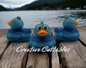 May include: Three blue and green rubber duck toys with textured surfaces are sitting on a wooden dock. The ducks are facing forward, with the middle duck in focus. The text "Creative Cuttables" is visible on the dock.