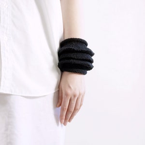 May include: A black knitted bracelet with a layered design. The bracelet is worn on a person's wrist.