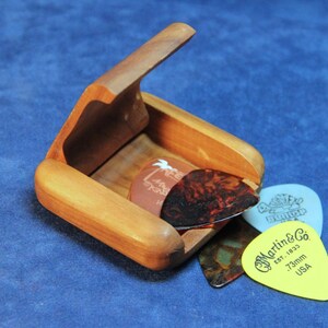 May include: A wooden box with a hinged lid, open to reveal four guitar picks. The picks are various colors and designs, including a white pick with a palm tree design, a brown pick with a black design, a blue pick with a white design, and a yellow pick with the text "Martin & Co. Est. 1833 73mm USA".