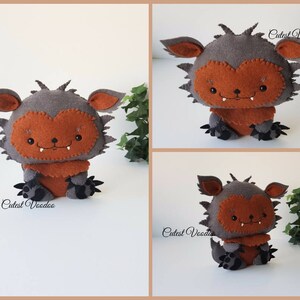 May include: A gray and orange felt monster plush toy with a toothy grin. The toy is sitting on a white surface with green leaves in the background. The text "Cutest Voodoo" is visible on the toy.