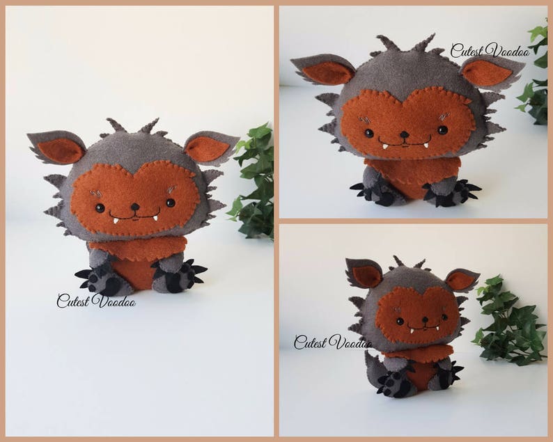 May include: A gray and orange felt monster plush toy with a toothy grin. The toy is sitting on a white surface with green leaves in the background. The text "Cutest Voodoo" is visible on the toy.