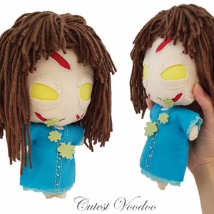 May include: A handmade plush toy doll with brown yarn hair, a white face with red and green details, and a blue dress. The doll is holding a green leaf in its hand.