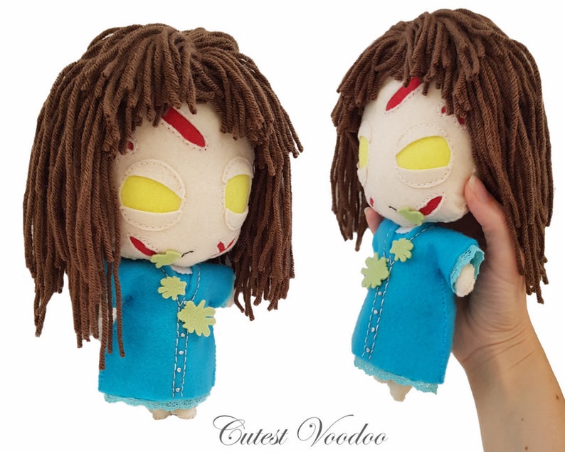 May include: A handmade plush toy doll with brown yarn hair, a white face with red and green details, and a blue dress. The doll is holding a green leaf in its hand.