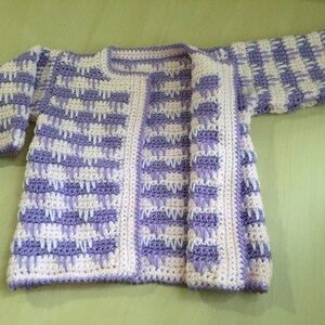 May include: A handmade crochet cardigan in a soft, open design. The sweater features a repeating pattern of lavender and white blocks. The edges and trim are a solid white, creating a clean border. The cardigan is displayed on a light green surface.