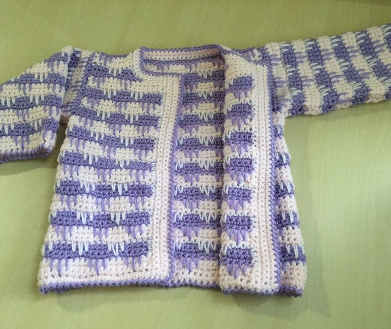 May include: A handmade crochet cardigan in a soft, open design. The sweater features a repeating pattern of lavender and white blocks. The edges and trim are a solid white, creating a clean border. The cardigan is displayed on a light green surface.