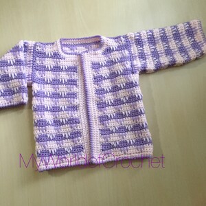 May include: A handmade crochet cardigan in a soft, pastel color palette of lavender and white. The open-front sweater features a textured stitch pattern and long sleeves. The text "MyWorldofCrochet" is visible at the bottom of the image.