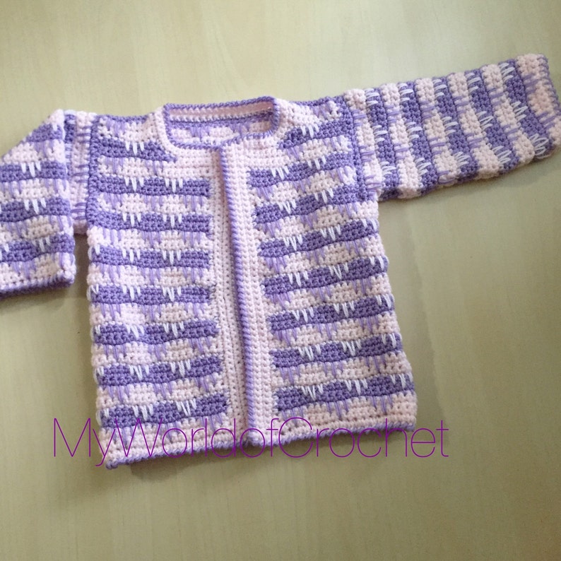 May include: A handmade crochet cardigan in a soft, pastel color palette of lavender and white. The open-front sweater features a textured stitch pattern and long sleeves. The text "MyWorldofCrochet" is visible at the bottom of the image.