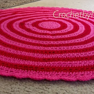 Op de afbeelding: Een handgemaakt, rond gehaakt kleed met concentrische cirkels in afwisselende tinten roze en rood. Het kleed heeft een geschulpte rand en ligt op een neutraal gekleurd tapijt. De tekst "CrochetbyNeelam.com" is zichtbaar.