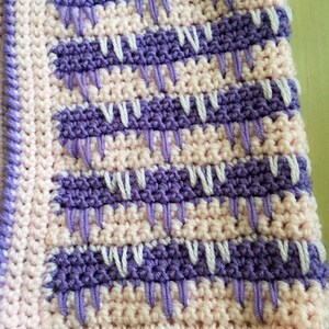 May include: A close-up of a crocheted blanket with a repeating pattern. The blanket features alternating rows of light pink and purple, with white and purple vertical stitches. The edges are finished with a purple border.