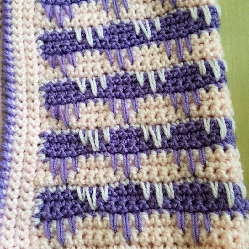 May include: A close-up of a crocheted blanket with a repeating pattern. The blanket features alternating rows of light pink and purple, with white and purple vertical stitches. The edges are finished with a purple border.