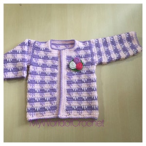 May include: A handmade crochet baby cardigan in shades of lavender and light pink. The sweater features a floral applique and a textured stitch pattern. The text "MyWorldofCrochet" is visible at the bottom. A charming and cozy garment.