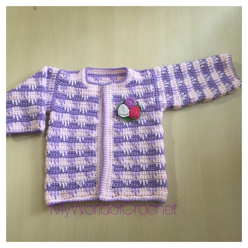 May include: A handmade crochet baby cardigan in shades of lavender and light pink. The sweater features a floral applique and a textured stitch pattern. The text "MyWorldofCrochet" is visible at the bottom. A charming and cozy garment.