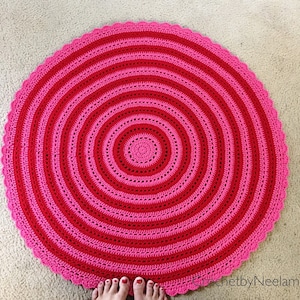 Op de afbeelding: Een handgemaakt, rond gehaakt vloerkleed in roze en roodtinten. Het kleed heeft concentrische cirkels in afwisselende kleuren, met een geschulpte rand. Het kleed ligt op een neutraal gekleurd tapijt.