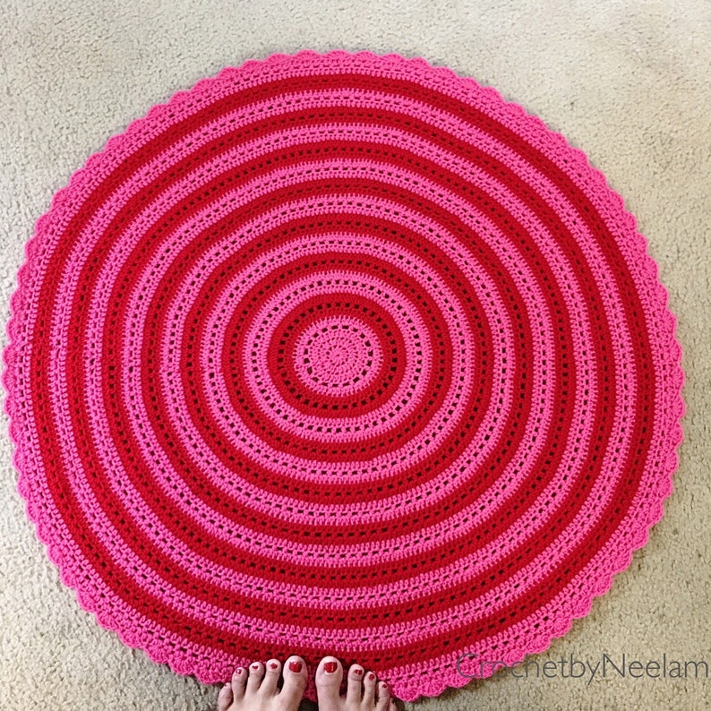 Op de afbeelding: Een handgemaakt, rond gehaakt vloerkleed in roze en roodtinten. Het kleed heeft concentrische cirkels in afwisselende kleuren, met een geschulpte rand. Het kleed ligt op een neutraal gekleurd tapijt.