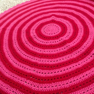 Op de afbeelding: Een handgemaakte, cirkelvormige gehaakte deken met concentrische ringen van afwisselend rood en roze. Het ontwerp van de deken cre&euml;ert een spiraaleffect, met een effen rood centrum en een reeks roze en rode banden. Het haakwerk is zichtbaar.