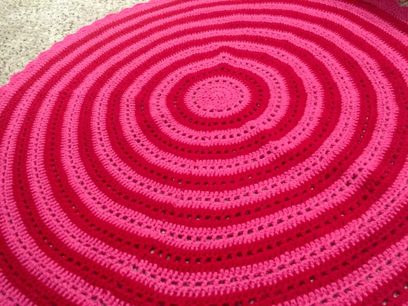 Op de afbeelding: Een handgemaakte, cirkelvormige gehaakte deken met concentrische ringen van afwisselend rood en roze. Het ontwerp van de deken cre&euml;ert een spiraaleffect, met een effen rood centrum en een reeks roze en rode banden. Het haakwerk is zichtbaar.
