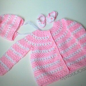 May include: A handmade baby set in pink and white crochet. The set includes a cardigan, bonnet, and booties. The cardigan has long sleeves and button closures. The bonnet has a ribbon tie. The booties have a small flower detail. The set is designed for a baby.