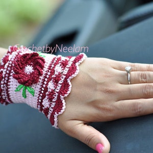 Puede incluir: Puño de muñeca de ganchillo en rosa y burdeos, con un diseño floral y detalles verdes. El puño tiene un borde festoneado y pequeñas cuentas blancas. El texto en la imagen dice "CrochetbyNeelam".