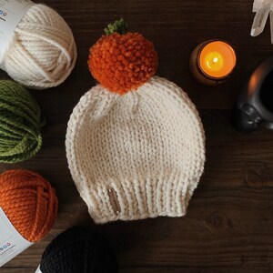 Puede incluir: Gorro de punto color crema con un pomp&oacute;n naranja y un tallo verde, que se asemeja a una calabaza. El gorro est&aacute; hecho a mano y se encuentra sobre una superficie de madera, rodeado de madejas de hilo de varios colores.