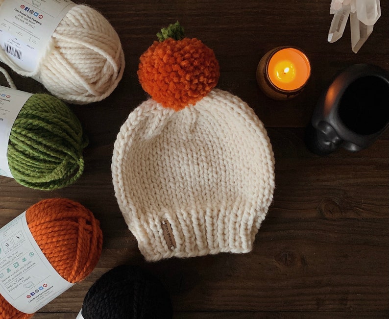 Puede incluir: Gorro de punto color crema con un pomp&oacute;n naranja y un tallo verde, que se asemeja a una calabaza. El gorro est&aacute; hecho a mano y se encuentra sobre una superficie de madera, rodeado de madejas de hilo de varios colores.