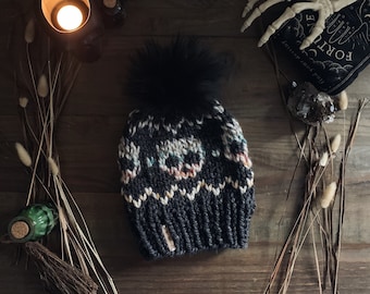 Op de afbeelding: Zwarte gebreide beanie met een wit en kleurrijk schedelpatroon en een zwarte pompon bovenop.