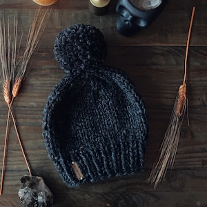 May include: A dark gray knitted beanie with a large pom pom on top. The beanie has a small leather tag with the word "Handmade" on it.