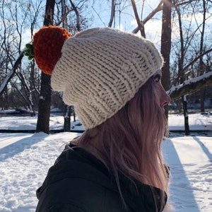 Puede incluir: Un gorro de punto grueso de color crema con un gran adorno en forma de zanahoria naranja y hojas verdes. El gorro se lleva al aire libre en un entorno nevado, con &aacute;rboles y luz solar en el fondo.
