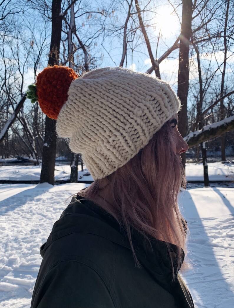 Puede incluir: Un gorro de punto grueso de color crema con un gran adorno en forma de zanahoria naranja y hojas verdes. El gorro se lleva al aire libre en un entorno nevado, con &aacute;rboles y luz solar en el fondo.