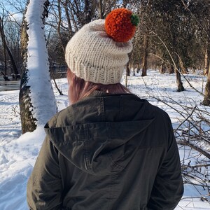 Puede incluir: Un gorro de punto color crema con un gran pomp&oacute;n naranja y un peque&ntilde;o acento verde. El gorro se usa al aire libre en un entorno nevado, con &aacute;rboles en el fondo. Es un accesorio de invierno.