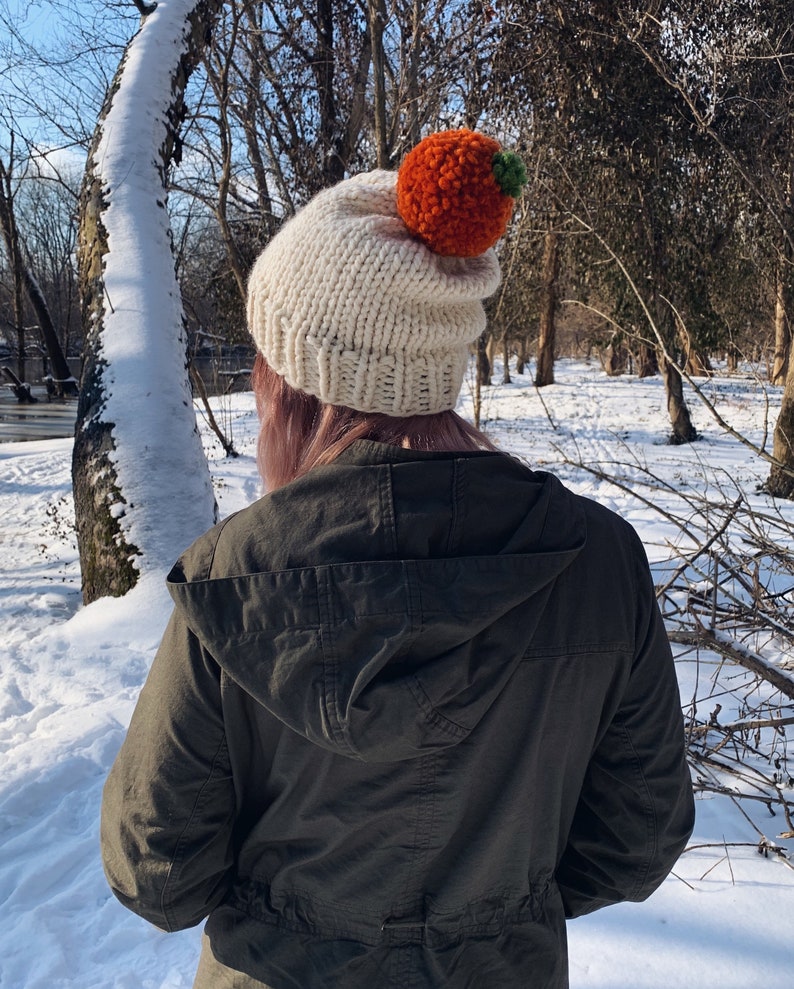 Puede incluir: Un gorro de punto color crema con un gran pomp&oacute;n naranja y un peque&ntilde;o acento verde. El gorro se usa al aire libre en un entorno nevado, con &aacute;rboles en el fondo. Es un accesorio de invierno.