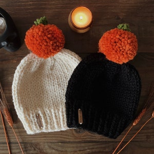 Puede incluir: Dos gorros de punto, uno crema y otro negro, cada uno coronado con un pomp&oacute;n naranja que se asemeja a una calabaza. Los gorros tienen una peque&ntilde;a etiqueta en el lateral. El fondo es una superficie de madera oscura con una vela encendida y elementos decorativos.