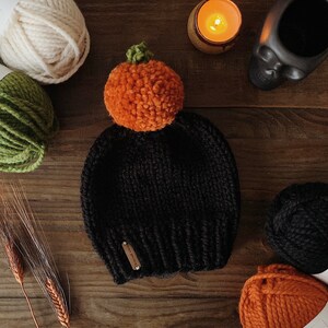 Puede incluir: Un gorro negro tejido con un pomp&oacute;n naranja con forma de calabaza y un tallo verde. El gorro tiene una peque&ntilde;a etiqueta rectangular. Madejas de lana en crema, verde, negro y naranja est&aacute;n dispuestas alrededor del sombrero.