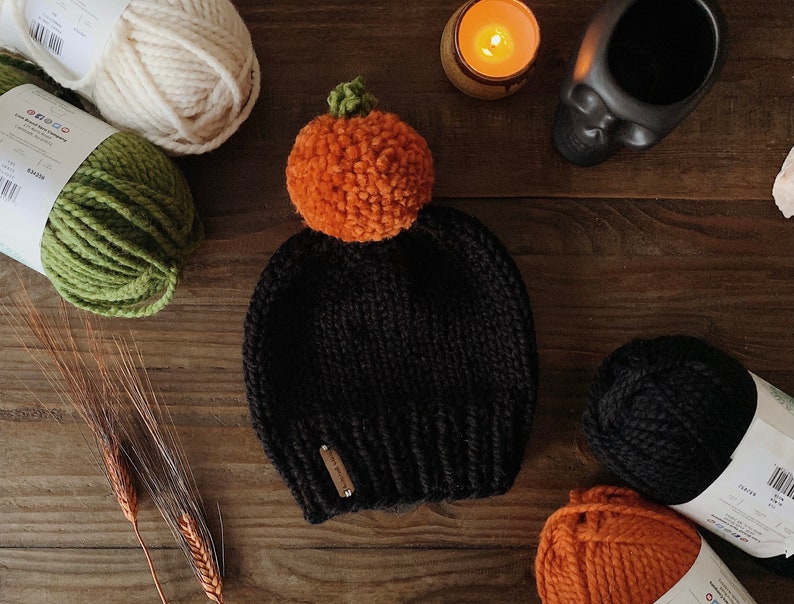 Puede incluir: Un gorro negro tejido con un pomp&oacute;n naranja con forma de calabaza y un tallo verde. El gorro tiene una peque&ntilde;a etiqueta rectangular. Madejas de lana en crema, verde, negro y naranja est&aacute;n dispuestas alrededor del sombrero.