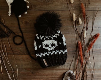 Op de afbeelding: Zwarte en witte gebreide beanie met een schedelpatroon en een zwarte nep bont pompon. De beanie heeft een bruin lederen label met het woord "Made" erop.
