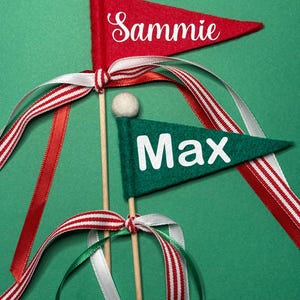 May include: Two felt pennant flags on wooden dowels. One is red with the name "Sammie" in white script, the other is green with "Max" in white block letters. Both have white pom-poms and are adorned with red, white, and green ribbons.