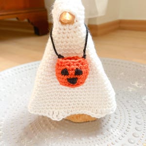 May include: A handmade Halloween decoration featuring a white crocheted ghost with a small orange pumpkin-shaped bag. The ghost has a glowing light inside. The pumpkin bag has a black face. The decoration is on a decorative plate.