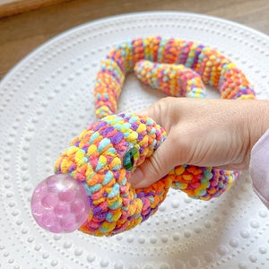 Könnte beinhalten: Ein farbenfrohes, gehäkeltes Schlangenspielzeug mit einer rosa Blasenstruktur am Kopf. Die Schlange ist aus weichem, plüschem Material gefertigt und perfekt zum Drücken und Spielen.