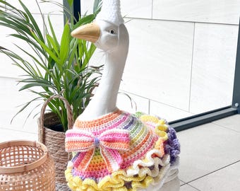 Könnte beinhalten: Eine weiße Gänsefigur, die ein gehäkeltes Outfit trägt, bestehend aus einem regenbogenfarbenen Oberteil mit einer großen Schleife an der Brust und einem gerüschten Rock. Die Gans trägt zudem einen weißen, kegelförmigen Partyhut mit einem Wollbommel.