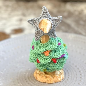 Op de afbeelding: Een handgemaakte gehaakte kerstboom met een grijze ster bovenop en kleurrijke lichtjes. De boom is groen met een textuurontwerp en staat op een goudkleurige basis. De boom wordt gepresenteerd op een grijze plaat.