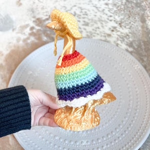 Puede incluir: Una caprichosa figura de ganso dorada con un atuendo de ganchillo hecho a mano. El atuendo incluye un vestido de arcoíris y un gorro amarillo. La figura se exhibe sobre un plato blanco texturizado.
