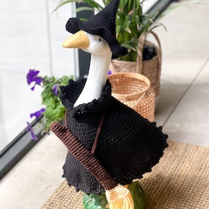 May include: A white goose figurine wearing a black crocheted witch hat and a matching black crocheted cape with a ruffled collar. A small brown crocheted broomstick with a light yellow straw-colored fringe is slung across the goose's body.