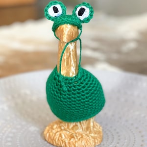 May include: A small wooden goose figurine wearing a green crocheted frog costume with white eyes. The goose is standing on a gray surface.