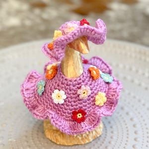 May include: A handcrafted, lavender-colored crochet duck figurine wearing a matching hat and dress, both adorned with colorful crocheted flowers. The duck is set against a neutral background, highlighting the intricate details of the handmade outfit.